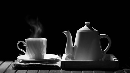 Aromatic hot tea or coffee in vintage white ceramic teacup with steam and teapot on wooden tabletop in black and white style, hot drink concept 