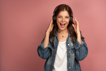 A girl in a jeans shirt stands on a pink background, clutching headphones and joyfully opening her mouth, looking at the camera.