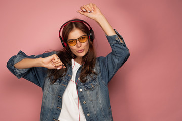A dark-haired girl in jeans stands on a pink background with headphones, dancing, looking at the camera.