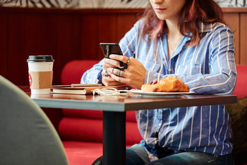 Close-up image of texting woman sitting at cafe table with cup of coffee and croissants on plate