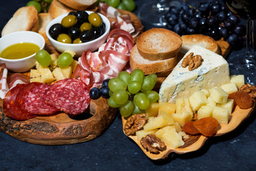 cheese platter on a wooden board, bread, fruit and cold cuts on dark background, closeup