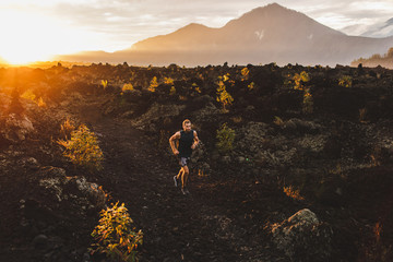 Young male athlete trail running in mountains at sunrise. Amazing black lava volcanic landscape of...
