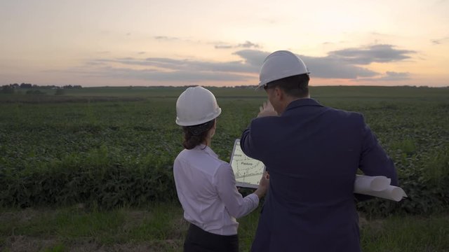 Man and woman discussing plans for agricultural and industrial development with blue prints and tablet and hard hats during golden hour sunset by a field of corn and beans