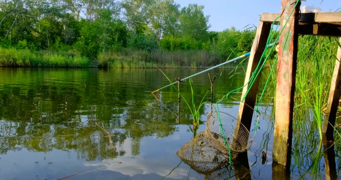 Fishing rod and float in the river in summer day. Angling tackle with a bobber and cage for caught fish. Nature background
