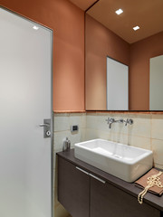 closeup of a ceramic counter top wash basin with its faucet on the wooden cabinet with a large mirror above on it, the walls are tinted of orange in the modern bathroom