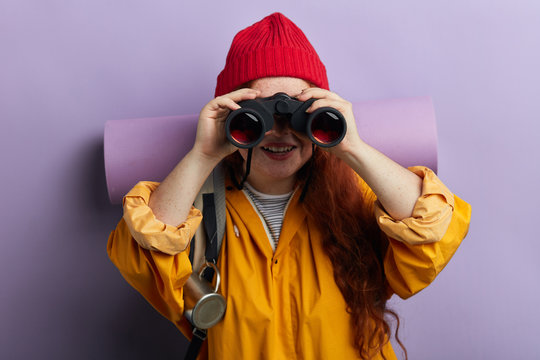 Beautiful young red-haired woman looking through binoculars.close up portrait, isolated blue background, studio shot, lifestyle, hobby, free time, spare time