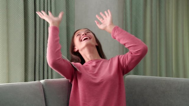smiling happy girl sits on grey sofa under money rain falling down from ceiling and catches banknotes