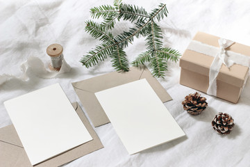 Christmas table, festive styled composition. Christmas greeting cards, envelopes mock-ups. Handmade gift box with silk ribbon, pine cones and fir tree branch on white linen background. Top view.