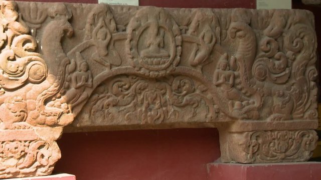 A daylight closeup shot of a stone lintel artifact on display inside the National Museum of Cambodia in Phnom Penh showing an intricate carved detailing depicting human figures, Buddha and more other