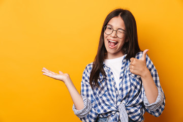 Happy pleased smiling young cute teenage girl in glasses posing isolated over yellow wall background showing copyspace make thumbs up.