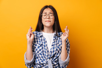 Nervous emotional young cute teenage girl in glasses posing isolated over yellow wall background showing hopeful please gesture.