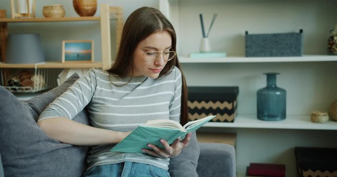 Smiling young woman student is reading interesting book indoors in cozy light studio apartment. Smart lady is wearing casual clothing and trendy glasses.
