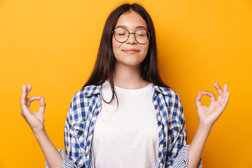 Pleased optimistic young cute teenage girl in glasses meditate isolated over yellow wall background.