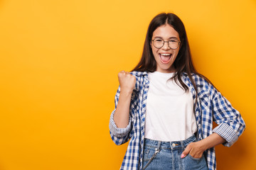 Emotional happy young cute teenage girl in glasses posing isolated over yellow wall background showing winner gesture.