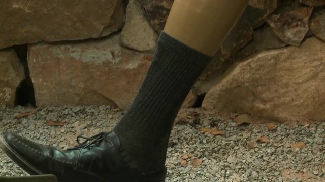 A closeup view and tilted downward shot of an army soldier veteran amputee wearing camouflage pants and showing off his plastic prosthetic leg wearing a black sock with matching black leather shoe at
