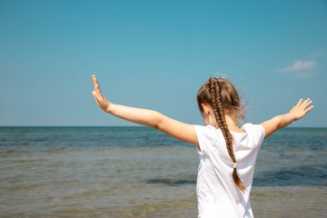 Fototapeta na wymiar a young girl in a white T-shirt with her hair combed into a long braid spread her arms sideways against the blue sea and blue sky. Copy space