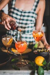 Young woman in checkered dress pouring Aperol to Aperol Spritz aperitif drink with orange in kitchen. Summer refreshing drink concept