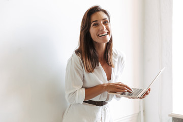 Fototapeta premium Cheerful smiling young business woman at workplace indoors near window using laptop computer.