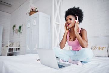 Fototapeta na wymiar Young african american woman sitting in bed while working on laptop and listen music on earphones.