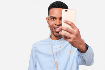 Image of happy African Amerfican young man holds modern smart phone, speaking online and blogging on cellphone. Handsome Afro male smiling and making self portrait on his device. People and technology
