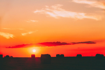 Summer Field Meadow With Hay Bales Silhouettes Under Sunset Sunbeams. Bright Sky And Dark Ground....
