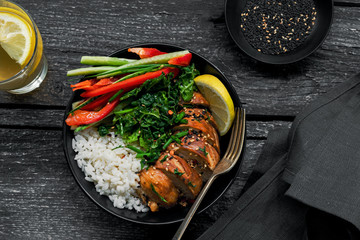 Buddha bowl meal with teriyaki chicken fillet, fresh vegetable salad, stew cabbage and basmati rice on a  table. Delicious dish in a black bowl with homemade lemonade. Top view shot.