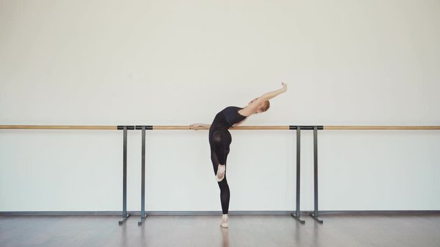 Side view tracking shot of graceful readhead young ballerina in black leotard practicing at barre in ballet studio