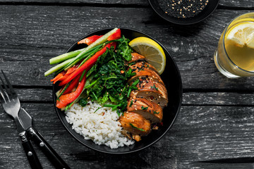 Buddha bowl meal with teriyaki chicken fillet, fresh vegetable salad, stew cabbage and basmati rice on a rustic table. Delicious dish in a black bowl with lemonade. Top view, directly above shot.