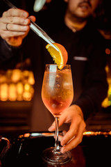 Elegant bartender is preparing pink cocktail holding orange chips at bar counter background.