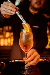 Elegant bartender is preparing pink cocktail holding orange chips at bar counter background.