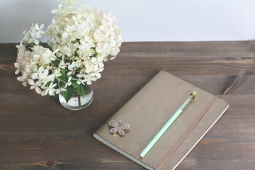 notepad with white hydrangea flowers in glasse vase on vintage wooden table. view from above