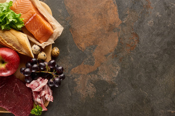 Top view of wooden cutting board with raw fish, meat, bacon, cheese, fruits, quail eggs, baguette and lettuce on dark brown marble surface with copy space
