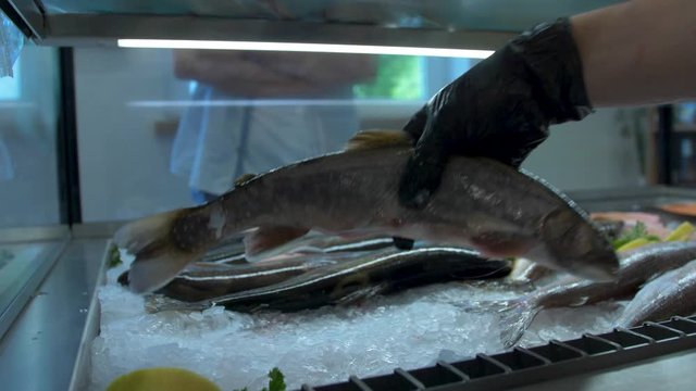 Medium close-up push-in shot of a hand wearing black gloves opening a store fridge and taking out a few pieces of Saibling char fish from over the ice with a customer waiting in the blurred background