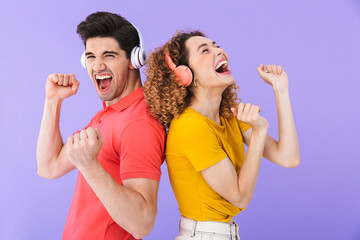Portrait of delighted couple laughing and listening to music with headphones standing back to back