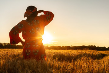 African woman in traditional clothes standing in a field of crops at sunset or sunrise - obrazy, fototapety, plakaty