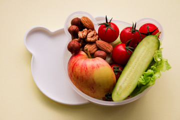 Children's lunch box shaped bear head with vegetarian food: vegetables, fruits, nuts on a light yellow background. Concept of healthy food, snack for children