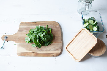 Wooden cutting board with spinach and cucumber and a knife and glass jars on white wooden background 