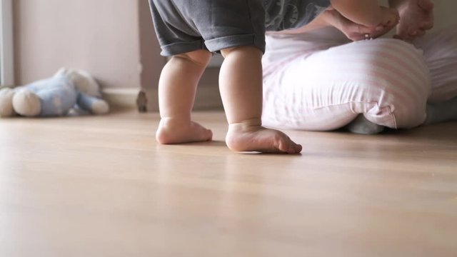 Close-up of baby feet learning to walk. 11 month toddler taking first steps at home, Mother looking at her child and happy smiling