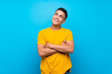 Young man with yellow shirt over isolated blue background looking up while smiling