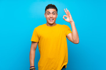 Young man with yellow shirt over isolated blue background surprised and showing ok sign