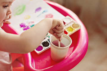 little girl of two years old is sitting in children's red chair and painting with watercolors