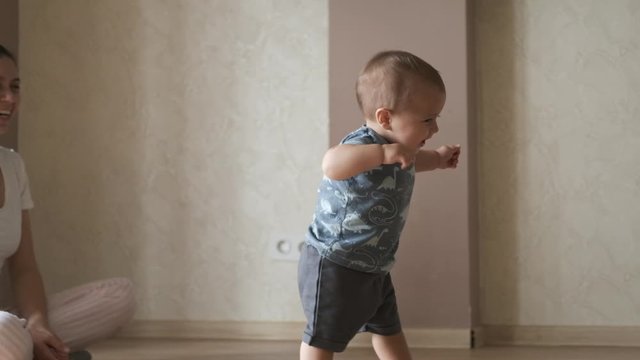 Cute baby doing the first steps and falls in slow motion at home near his mother. Smiles and feels happy