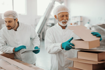 Fototapeta na wymiar Caucasian working senior in white uniform putting and preparing boxes while smiling supervisor holding tablet and checking boxes. Food plant interior.