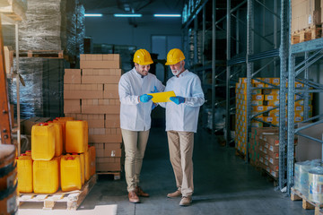 Two warehouse employees in white uniforms and with yellow helmets on heads standing and looking at documentation. All around are boxes with goods.