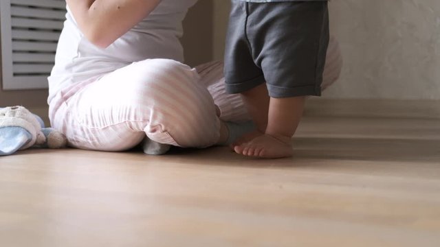 Close-up of baby feet learning to walk. 11 month toddler taking first steps at home, Mother looking at her child and happy smiling