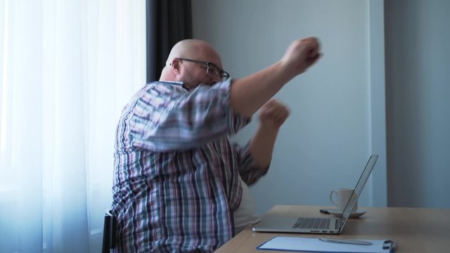 Happy funny fat bald man in glasses rejoices and emotionally expressively dances while sitting at table in office in front of laptop