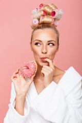 Portrait of caucasian young housewife in housecoat with hair curlers smiling while eating sweet...