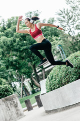 Full-length portrait of young woman trail runner jumping while having workout in city park. Vertical shot. Dutch angle