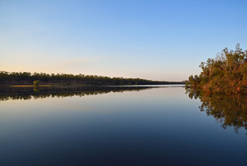 Stillness of calm river waters with late afternoon sun highlighting the trees and reflecting blue sky.