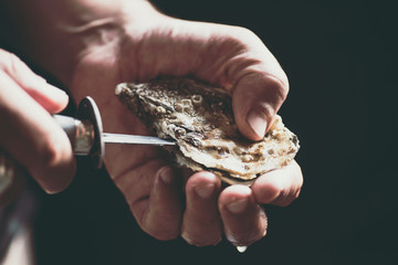 A man with a knife opens a fresh oyster, a drop of water on his hand. Dark rustic background,...